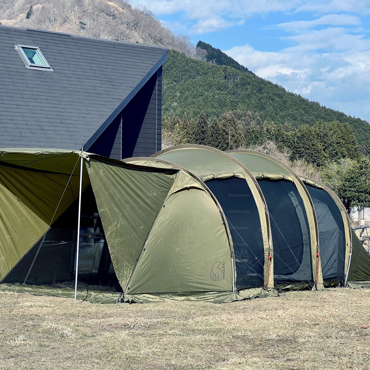 Green camping tent with attached porch in a scenic outdoor setting with mountains and a building.