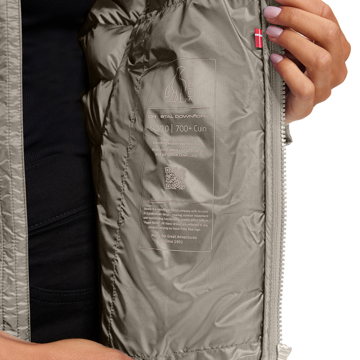 Close-up of the interior lining of a puffer jacket, showing a silver-gray insulated pocket with a printed product label and a small red flag tag, with a person's hand holding the zipper.