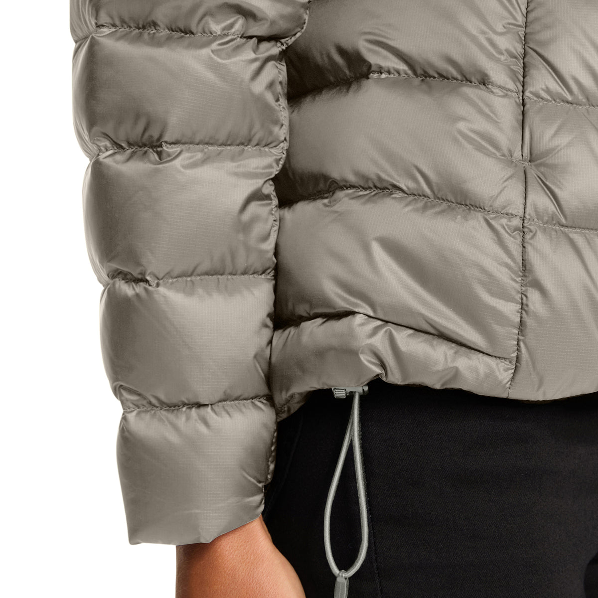Close-up of a person wearing a cropped, quilted puffer jacket in a taupe-gray color, paired with black pants. The jacket features horizontal baffles and a drawstring hem.