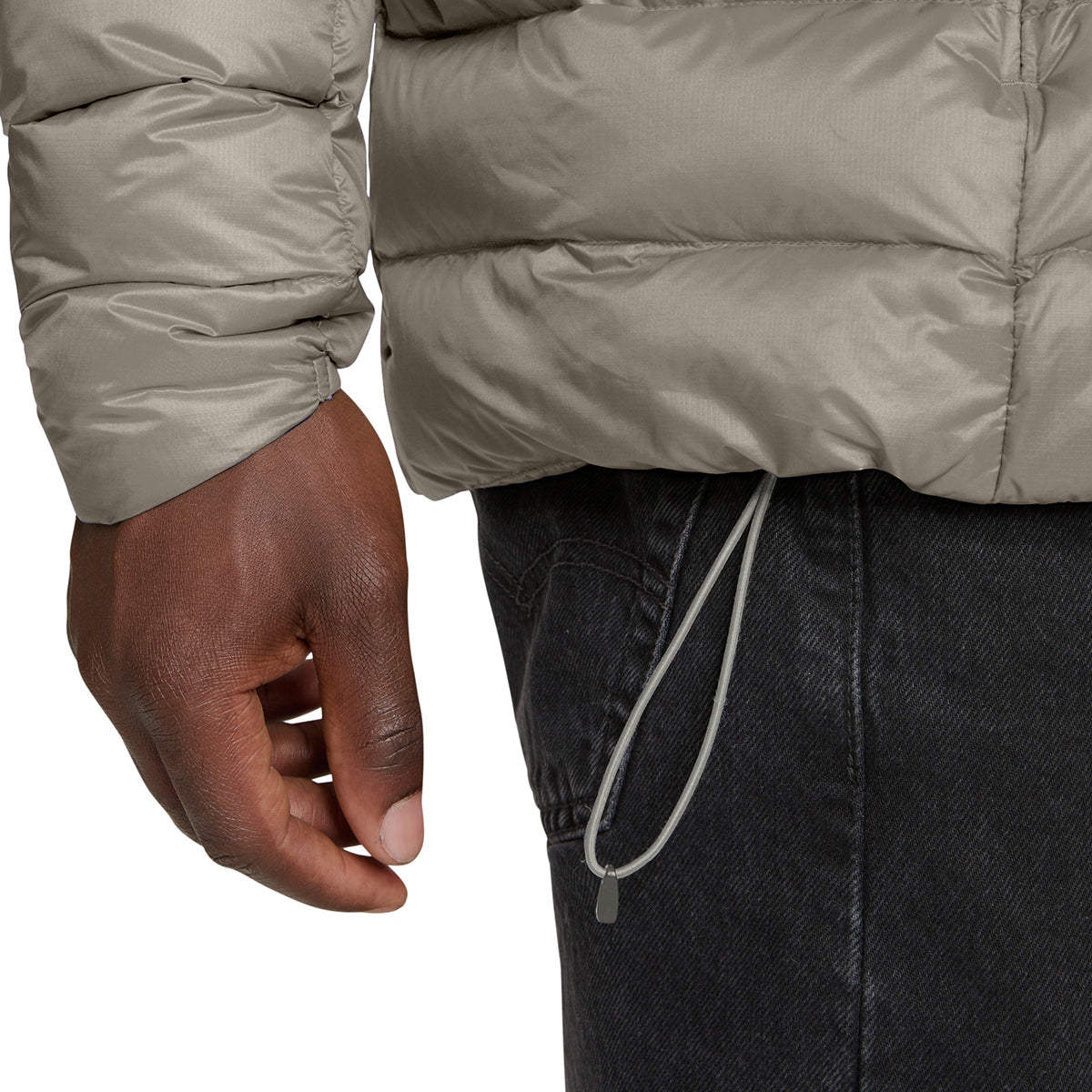 Close-up of a person wearing a light taupe puffer jacket and dark charcoal jeans, showing the sleeve cuff, hand, and drawstring waist.