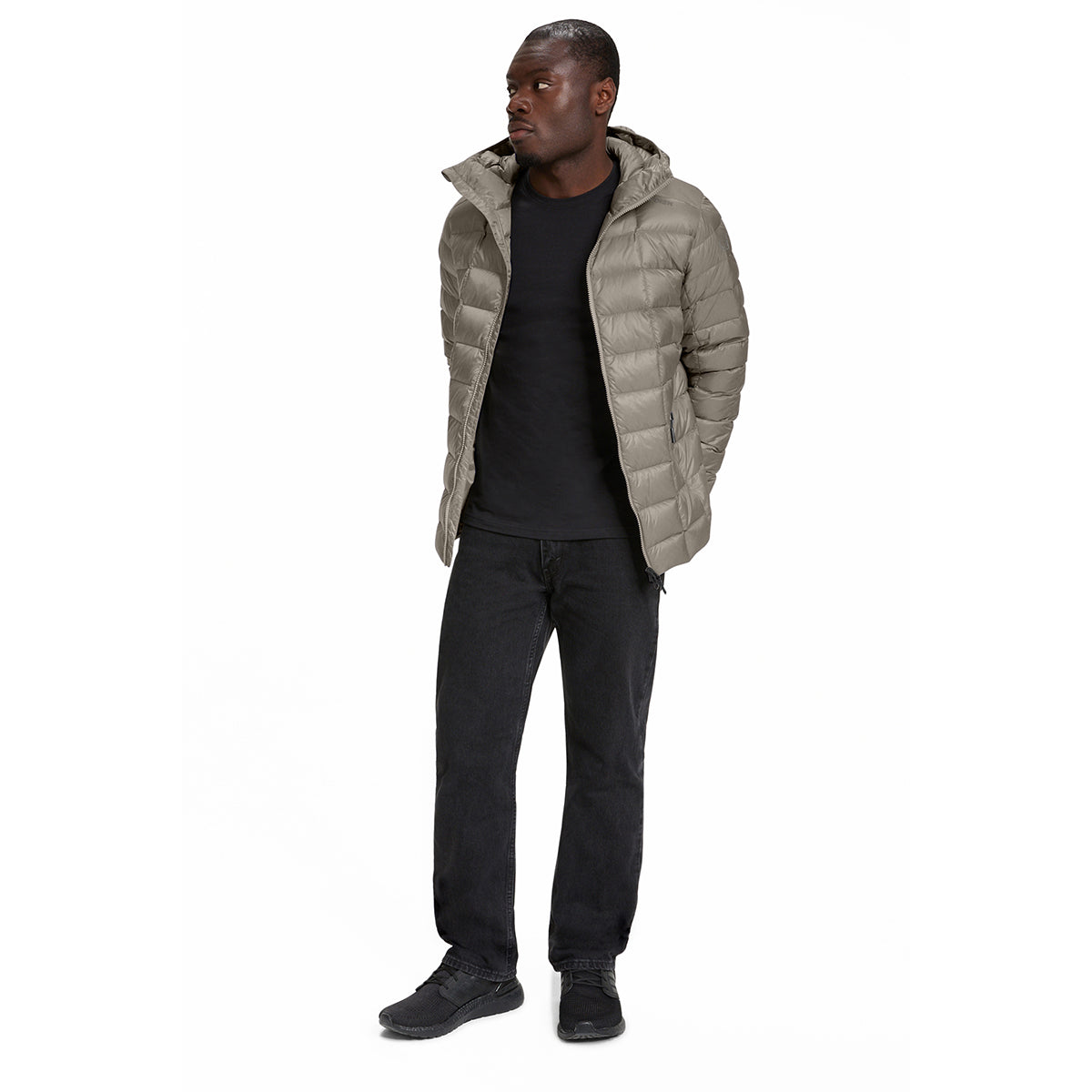 A man wearing a light taupe-beige hooded puffer jacket over a black t-shirt, black jeans, and black sneakers, standing against a solid white background.