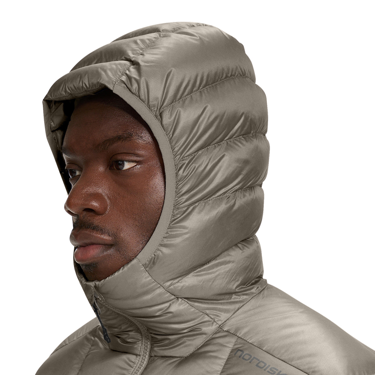 A close-up side profile of a man wearing a hooded puffer jacket in a light taupe-gray color, with the brand name 'nordisk' visible on the chest, against a solid white background.