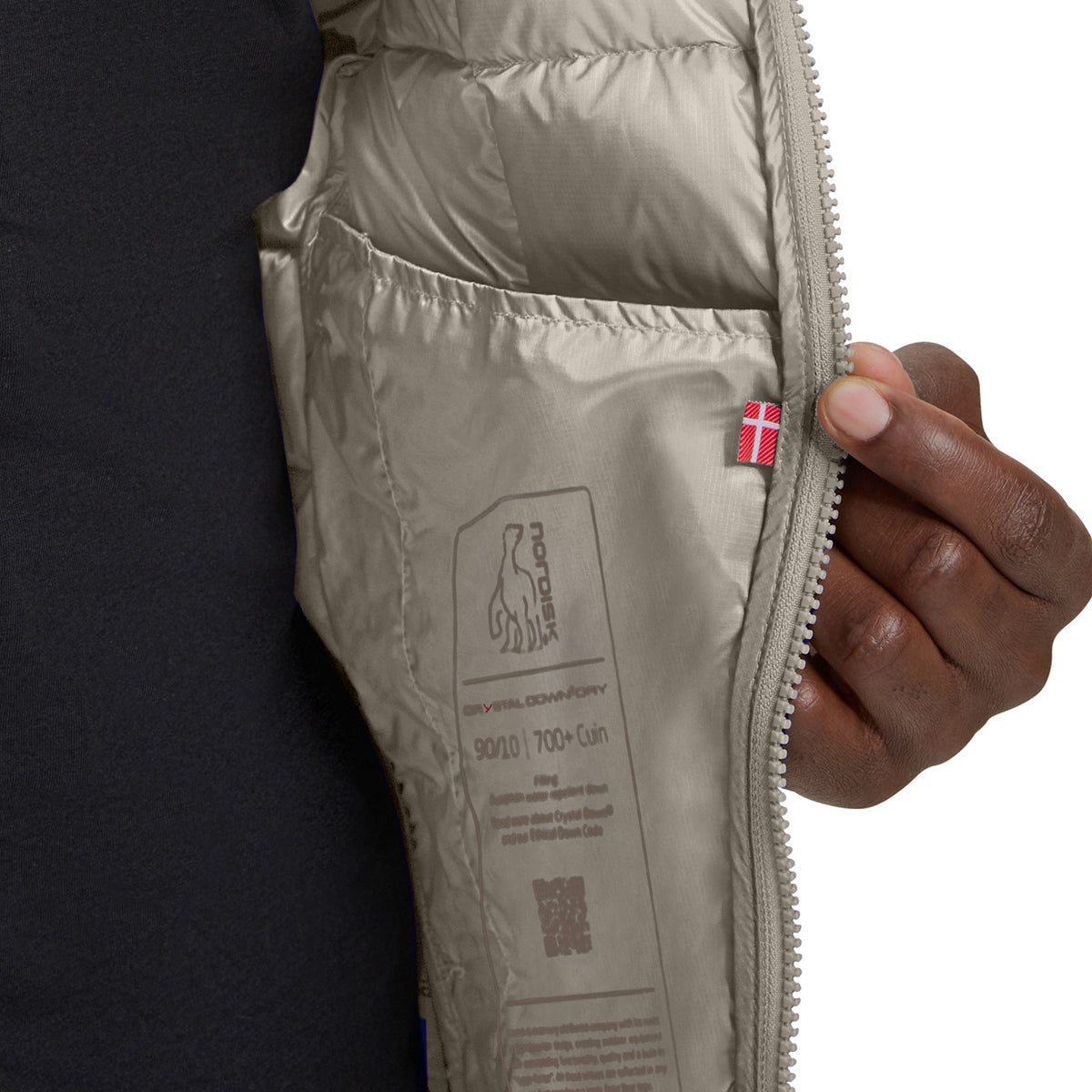 Close-up of a person's hand holding open a beige puffer jacket, showing the interior lining with a printed label and a small red and white flag tab near the zipper.