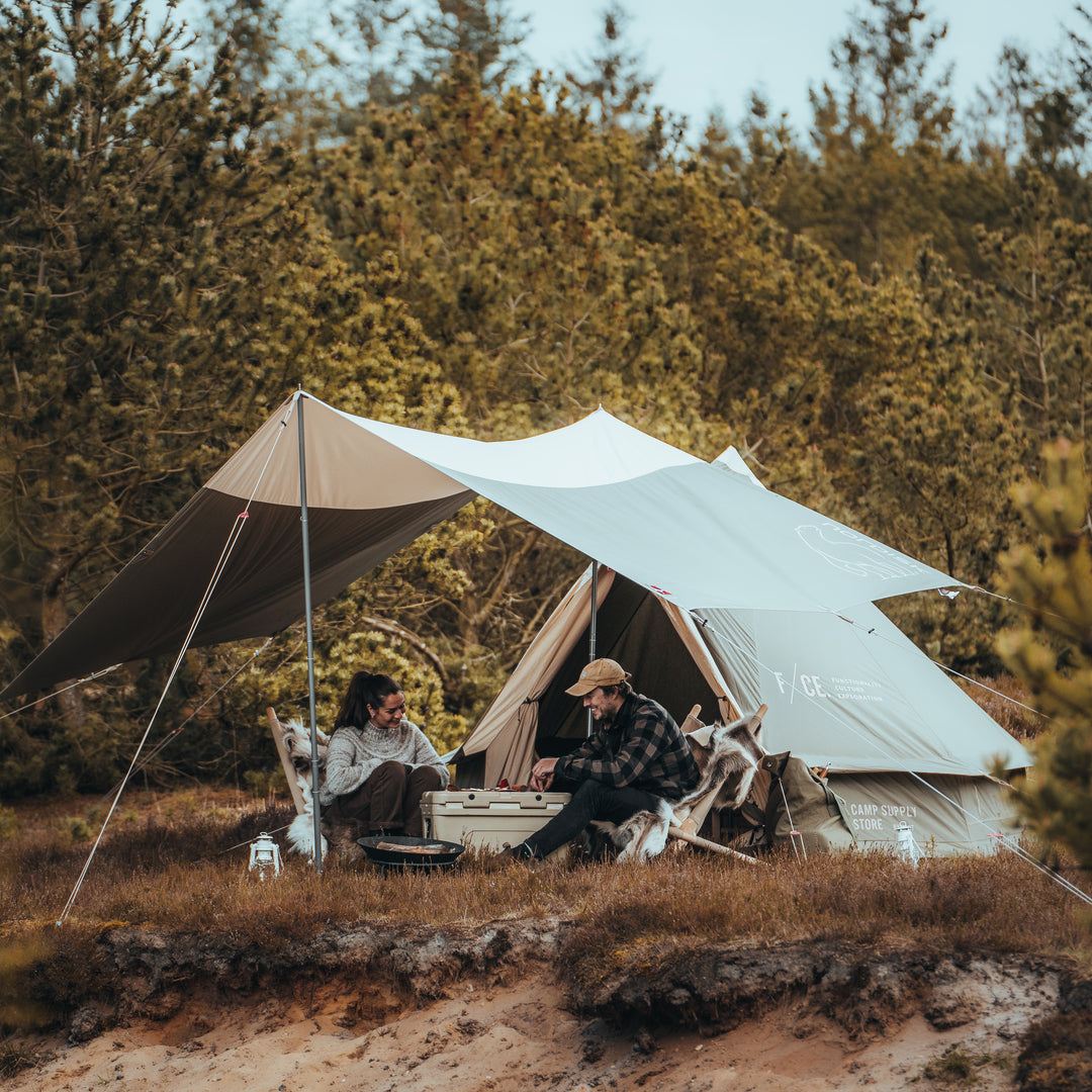 Two people camping outdoors with a tent and tarp in a forest setting.