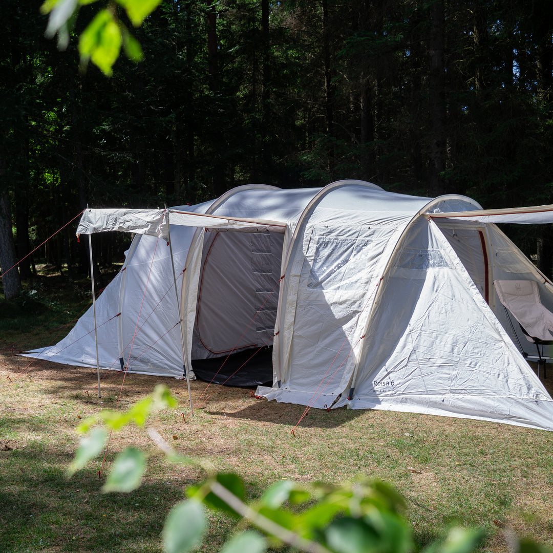 A large white and gray dome camping tent with an attached side awning, pitched on grass in a wooded forest setting.